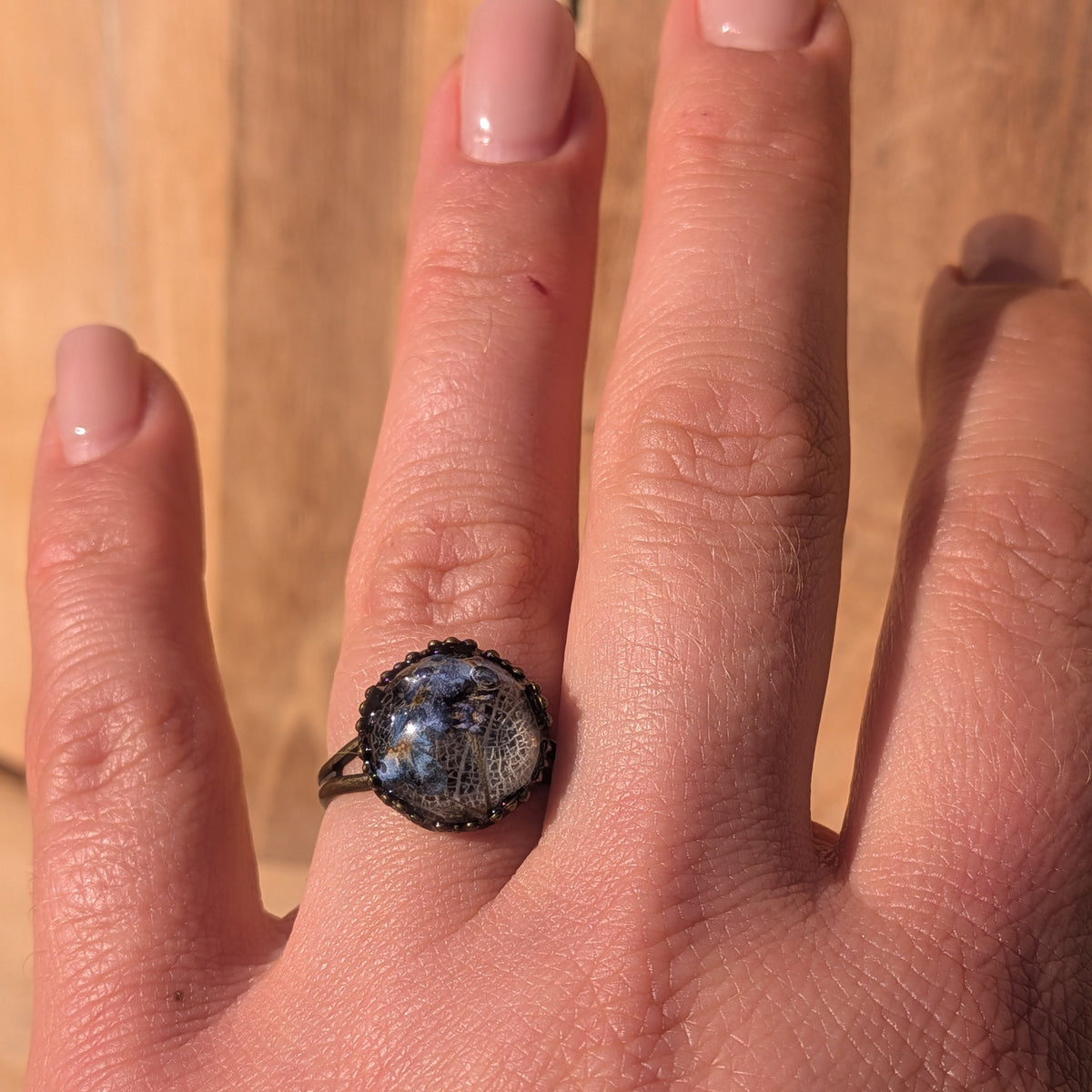 Blue forget-me-not and white skeleton leaf resin ring worn on an outstretched hand against a wooden background, EL-Creations