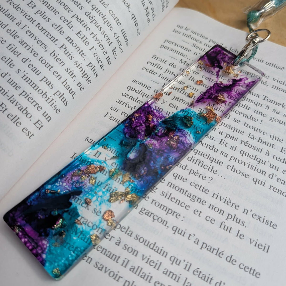 Full view of handmade cosmic resin bookmark with teal tassel on open book page – EL-Creations