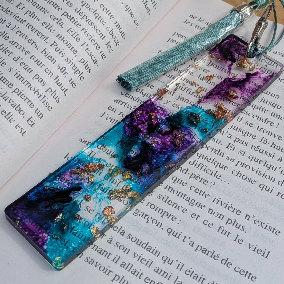 Handmade teal and purple resin bookmark lying across open French book pages – EL-Creations