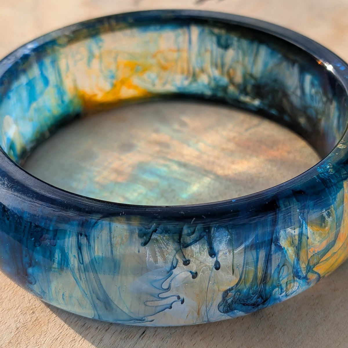Handmade deep blue and teal resin bangle with gold swirls lying on wooden surface – EL-Creations