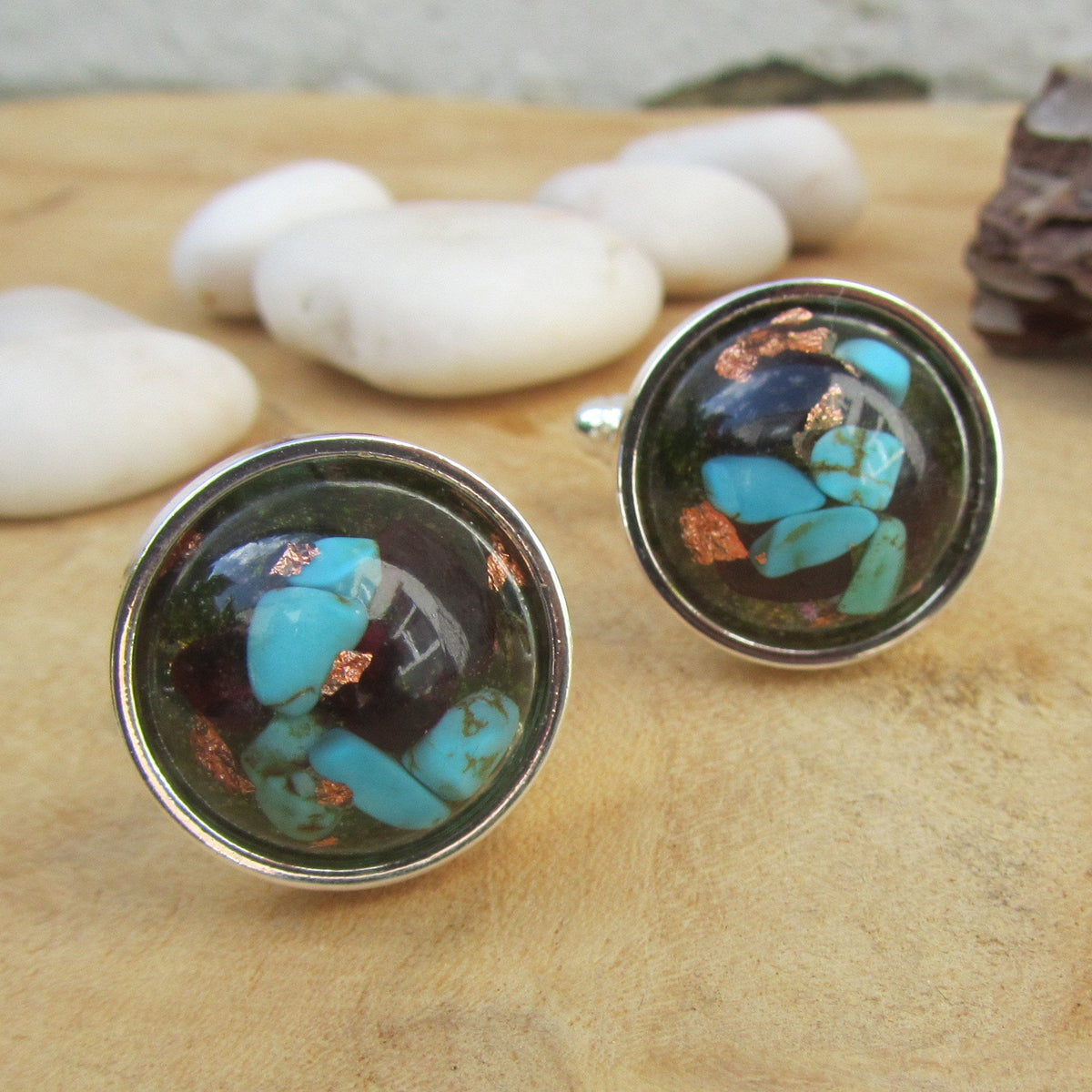 Pair of cufflinks with turquoise and copper design on a wooden surface with pebbles.