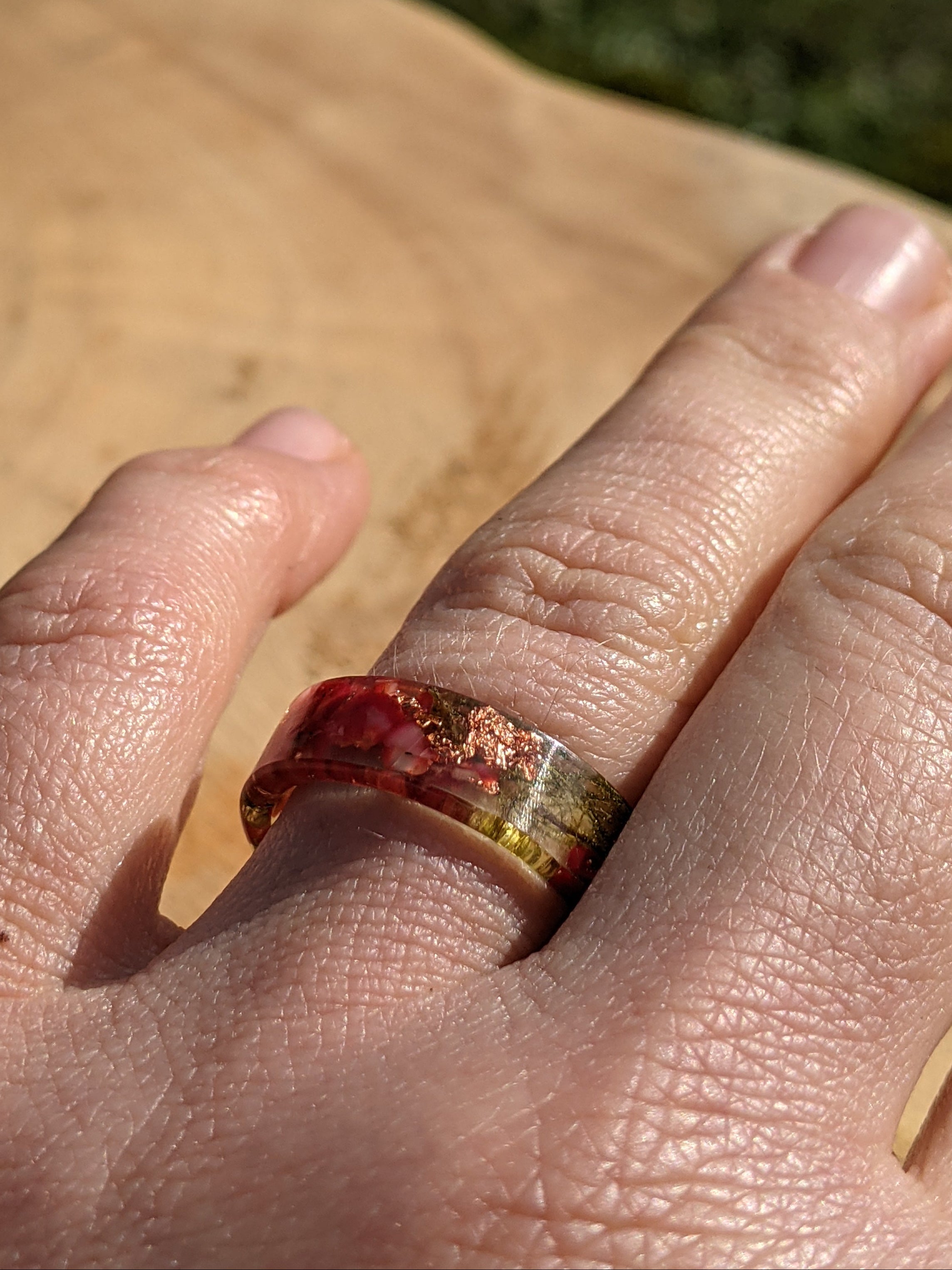 Hand wearing a ring with a multicolored stone on a wooden surface with grass in the background