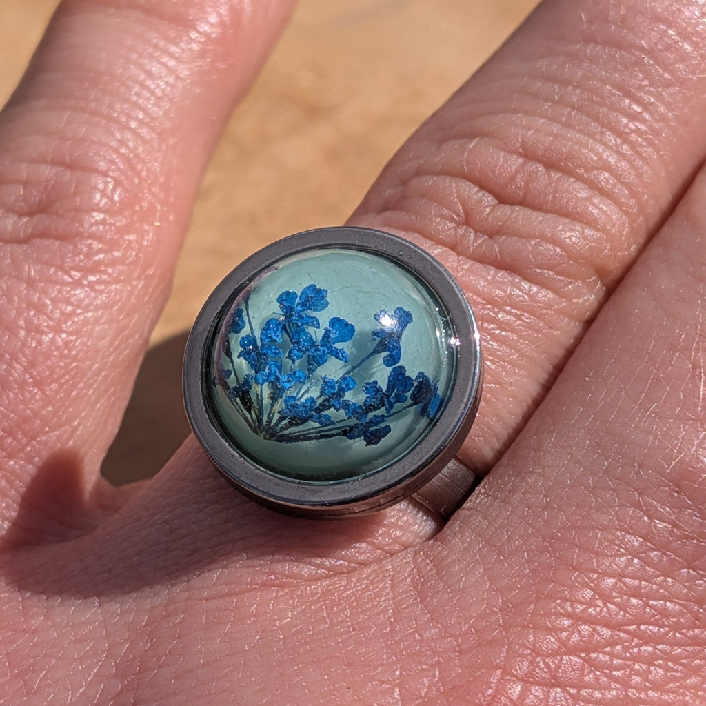 Blue botanical resin ring worn on finger, close-up of teal cabochon with pressed flowers in steel bezel – EL-Creations