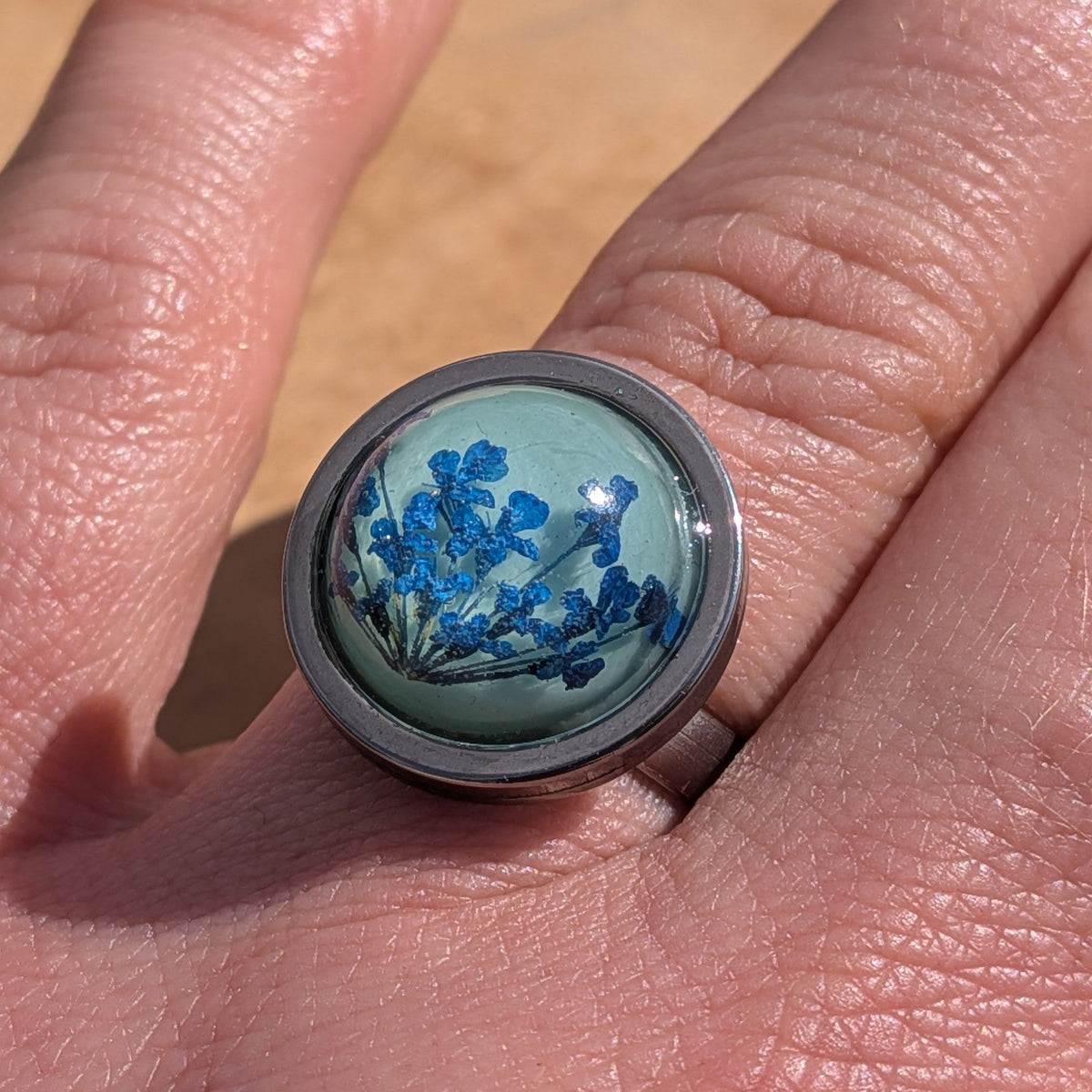 Blue botanical resin ring worn on finger, close-up of teal cabochon with pressed flowers in steel bezel – EL-Creations