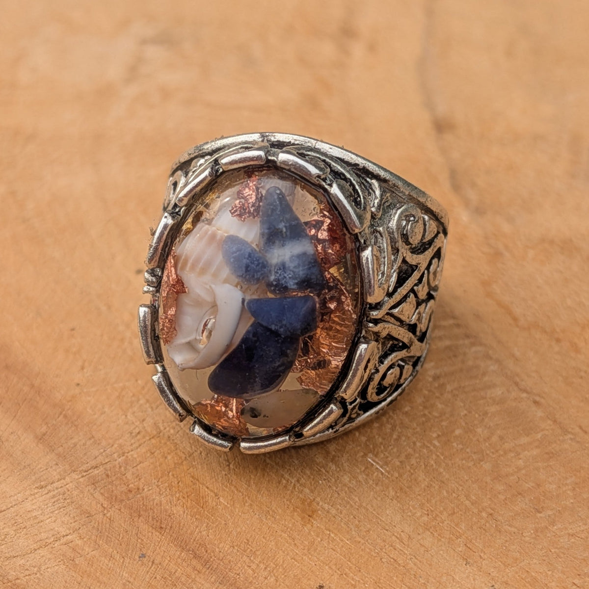 Clear resin oval ring with sodalite and copper flakes in baroque filigree band side view on wood – EL-Creations