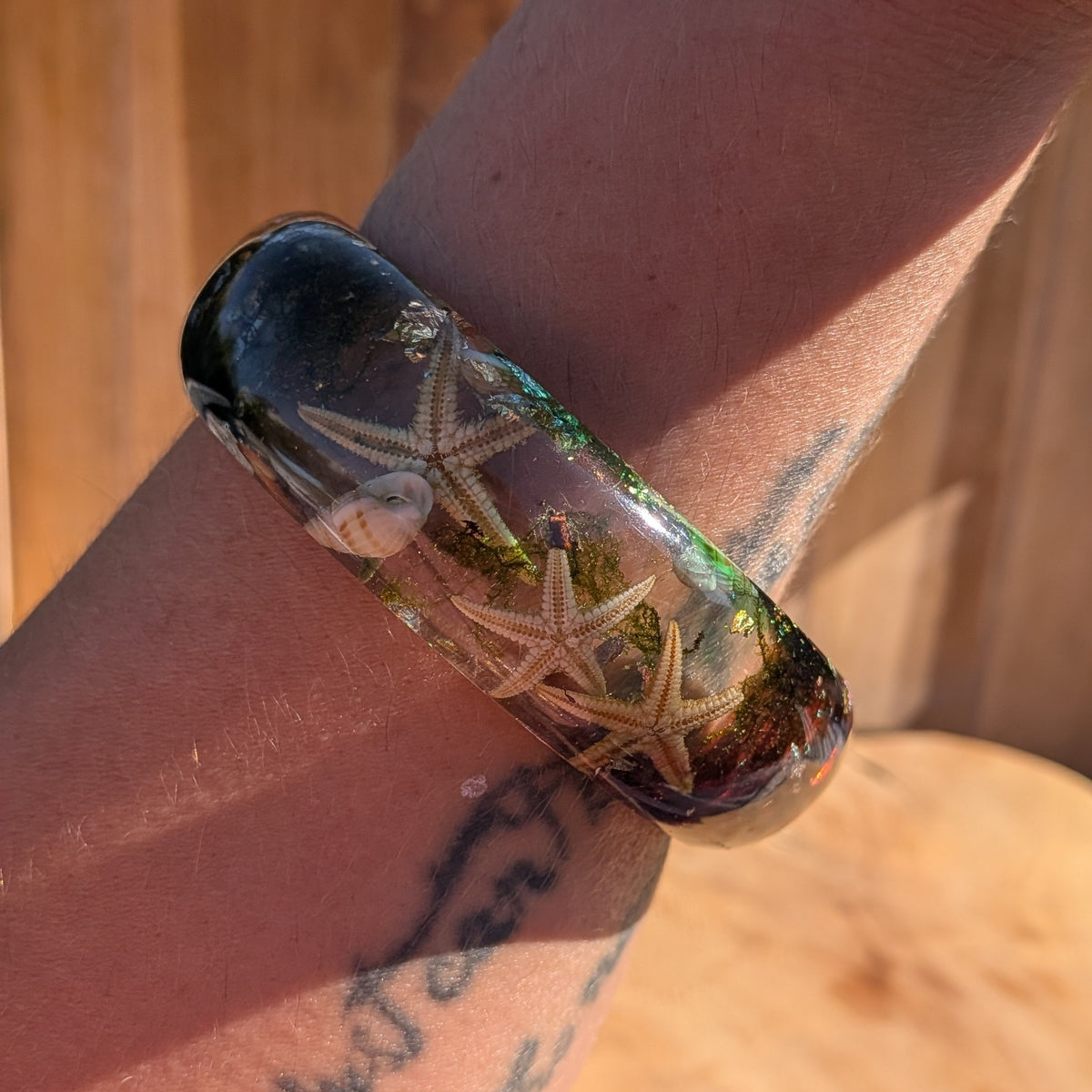 A close-up of a person's wrist wearing a thick, glossy resin cuff bracelet filled with starfish, shells, and green seaweed, set against a warm wooden background.