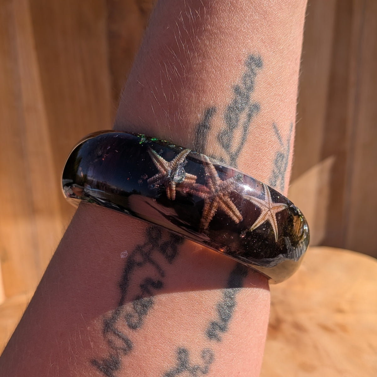 A close-up of a person's tattooed wrist wearing a glossy black bangle bracelet with three starfish inlay.