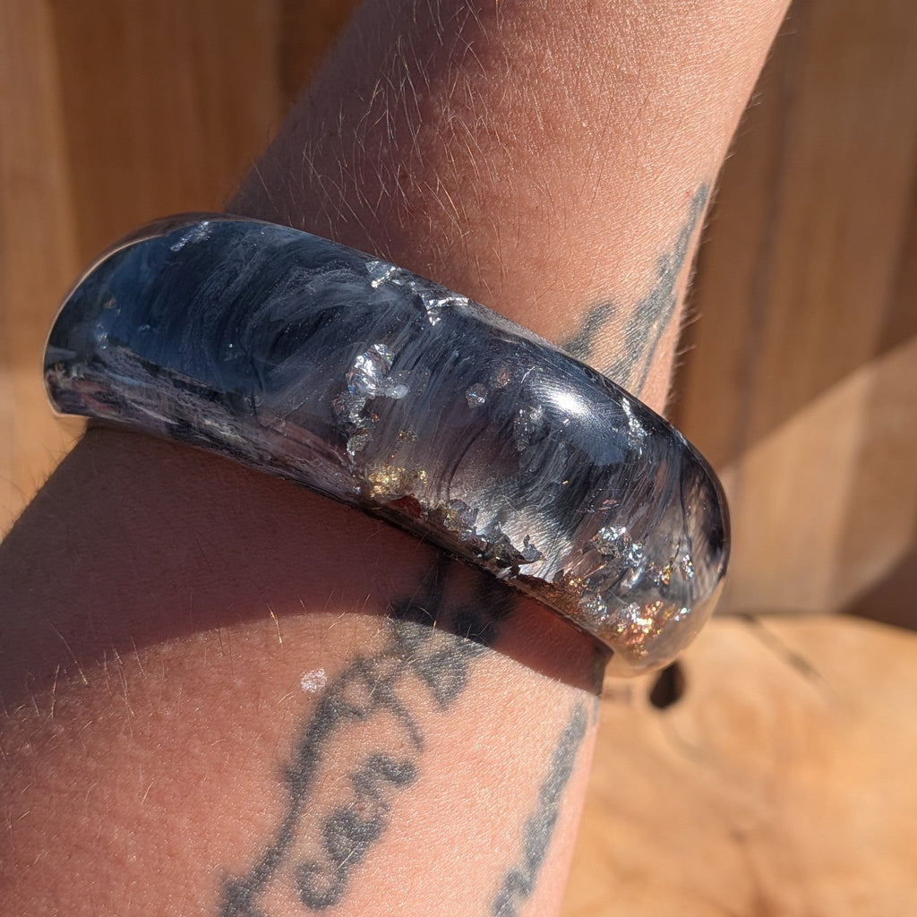 Chunky resin bangle black and white pigment swirls with gold copper silver flakes angled view on wood – EL-Creations