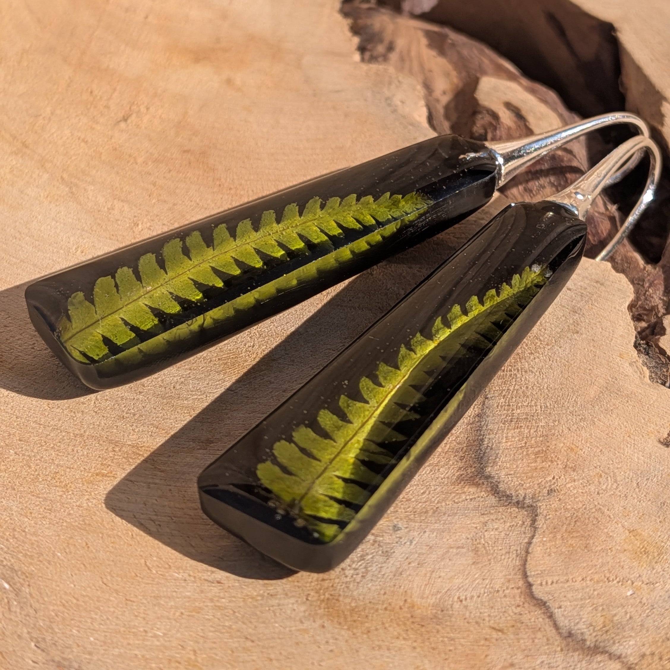 Black resin dangle earrings with fern – hanging against wooden wall in sunlight – EL-Creations