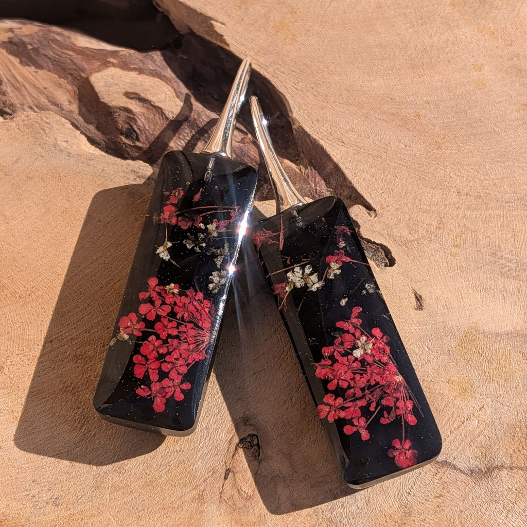 Black resin earrings with dried red flowers – leaning against driftwood in sunlight – EL-Creations