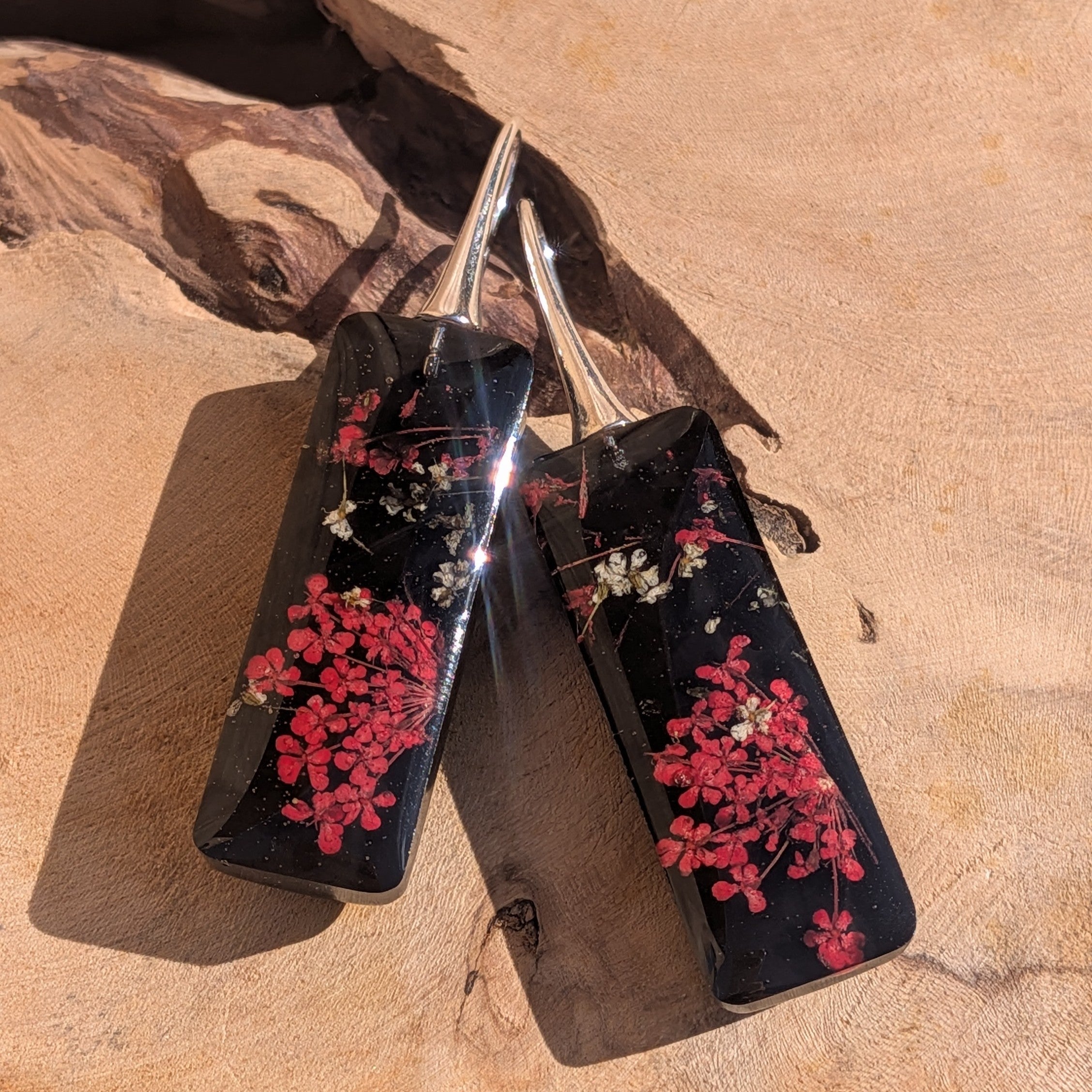 Black resin earrings with dried red flowers – leaning against driftwood in sunlight – EL-Creations