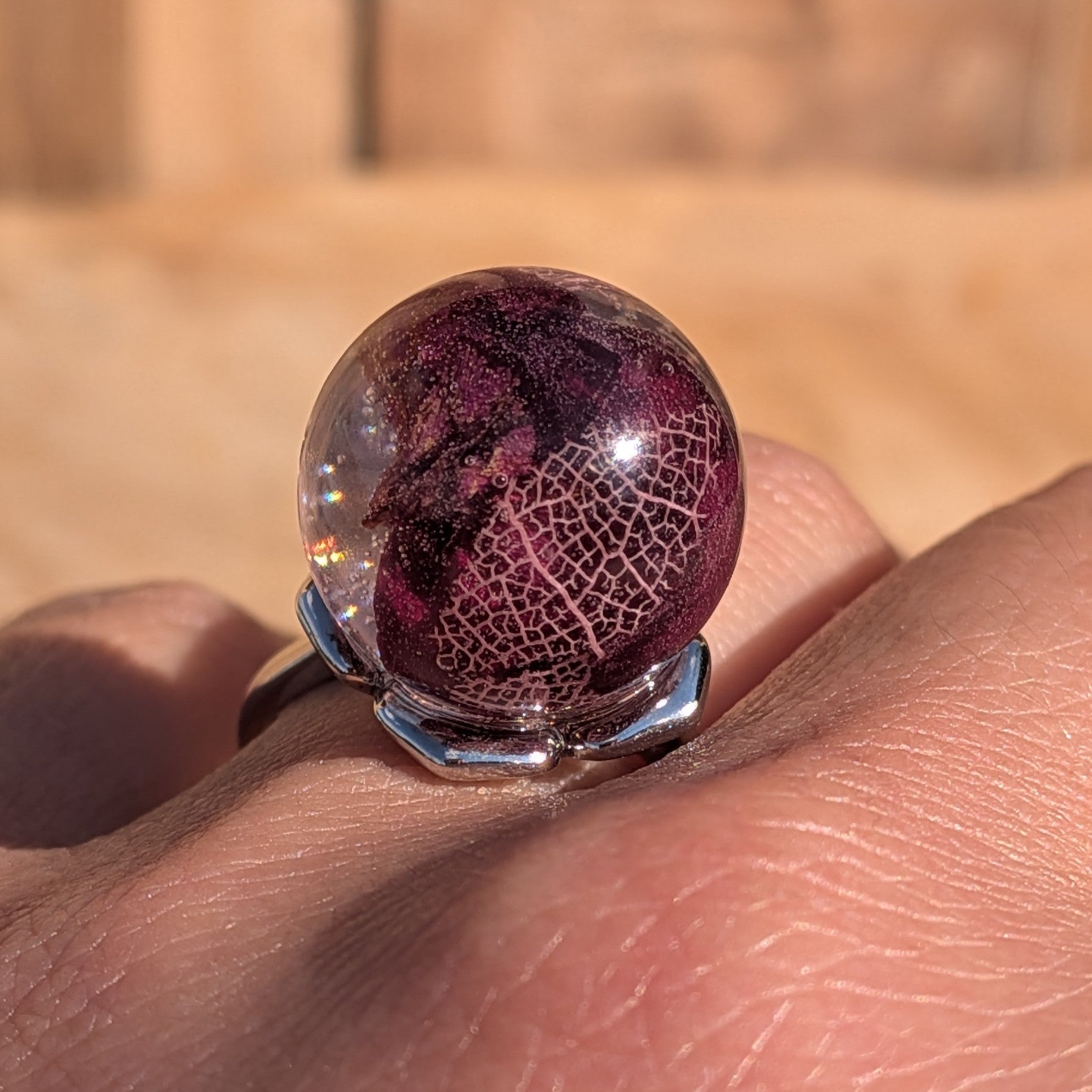 Dark burgundy resin ring with rose petals and skeleton leaf worn on finger – wooden background – EL-Creations