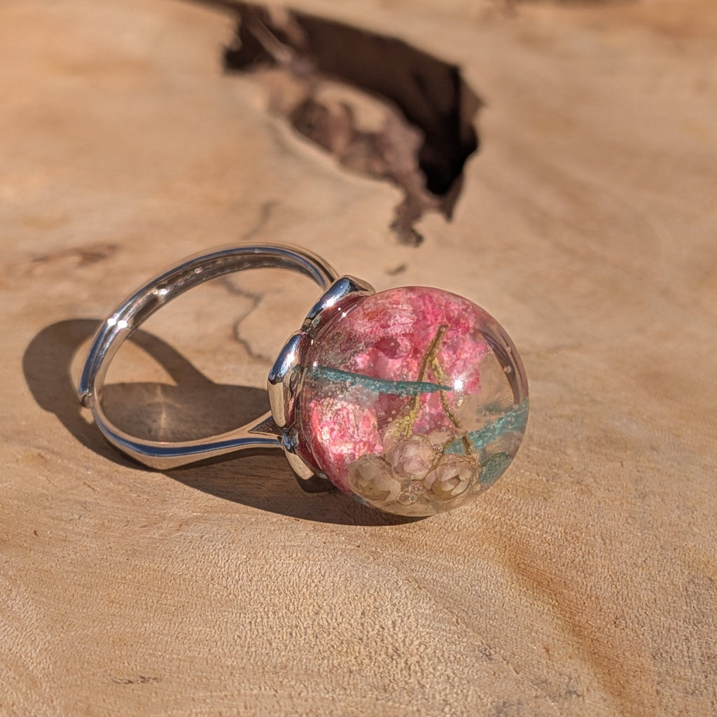 Handmade resin globe ring lying on wood surface showing pink wildflower and teal skeleton leaf – EL-Creations