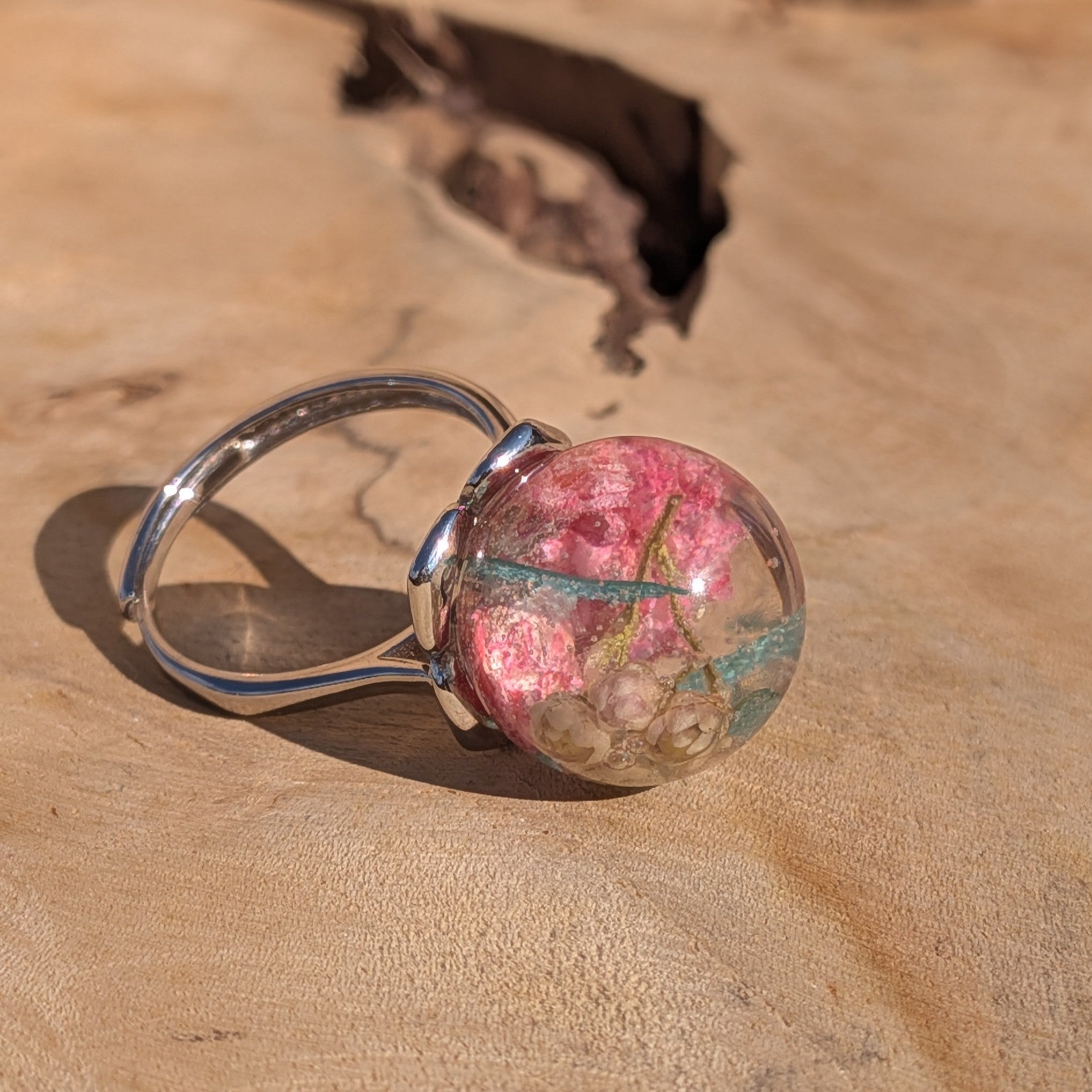 Handmade resin globe ring lying on wood surface showing pink wildflower and teal skeleton leaf – EL-Creations
