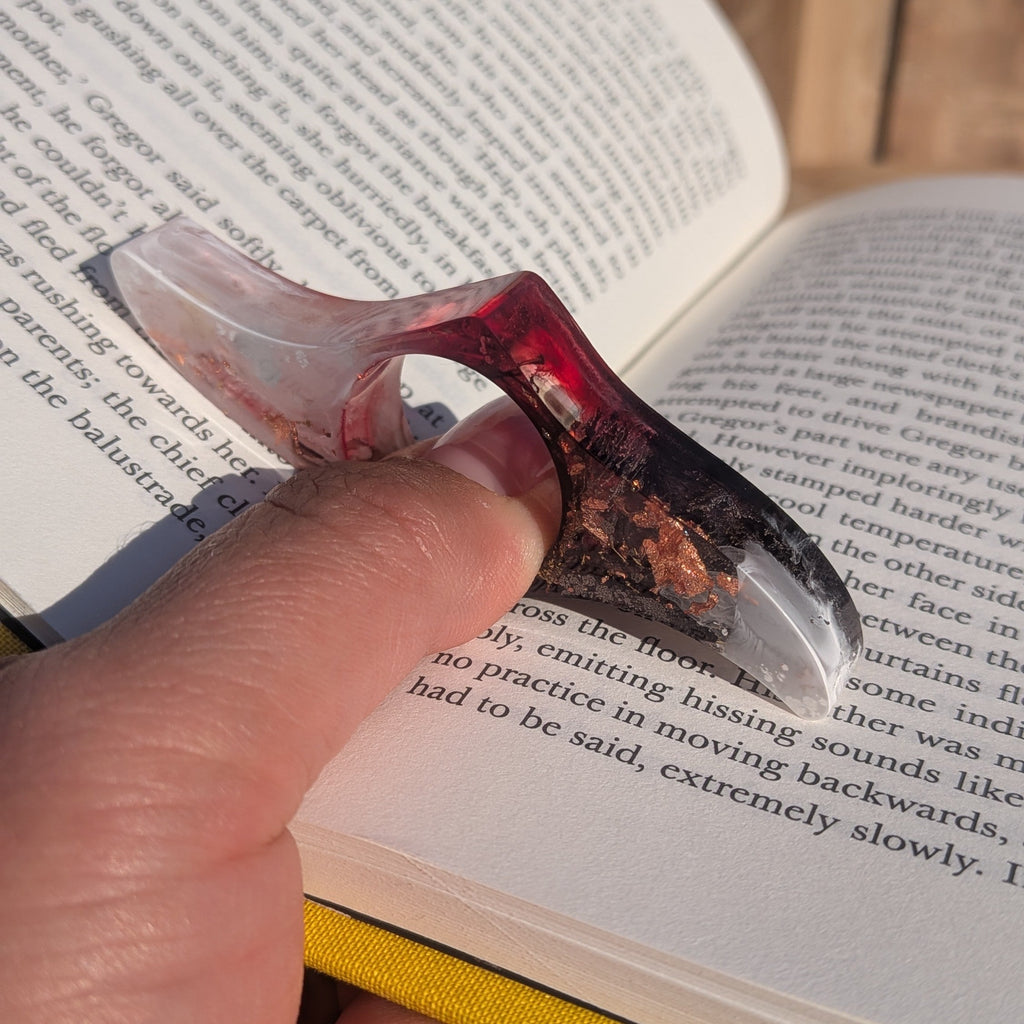 Resin reading ring with copper flakes in use on thumb over open book – close-up view – EL-Creations