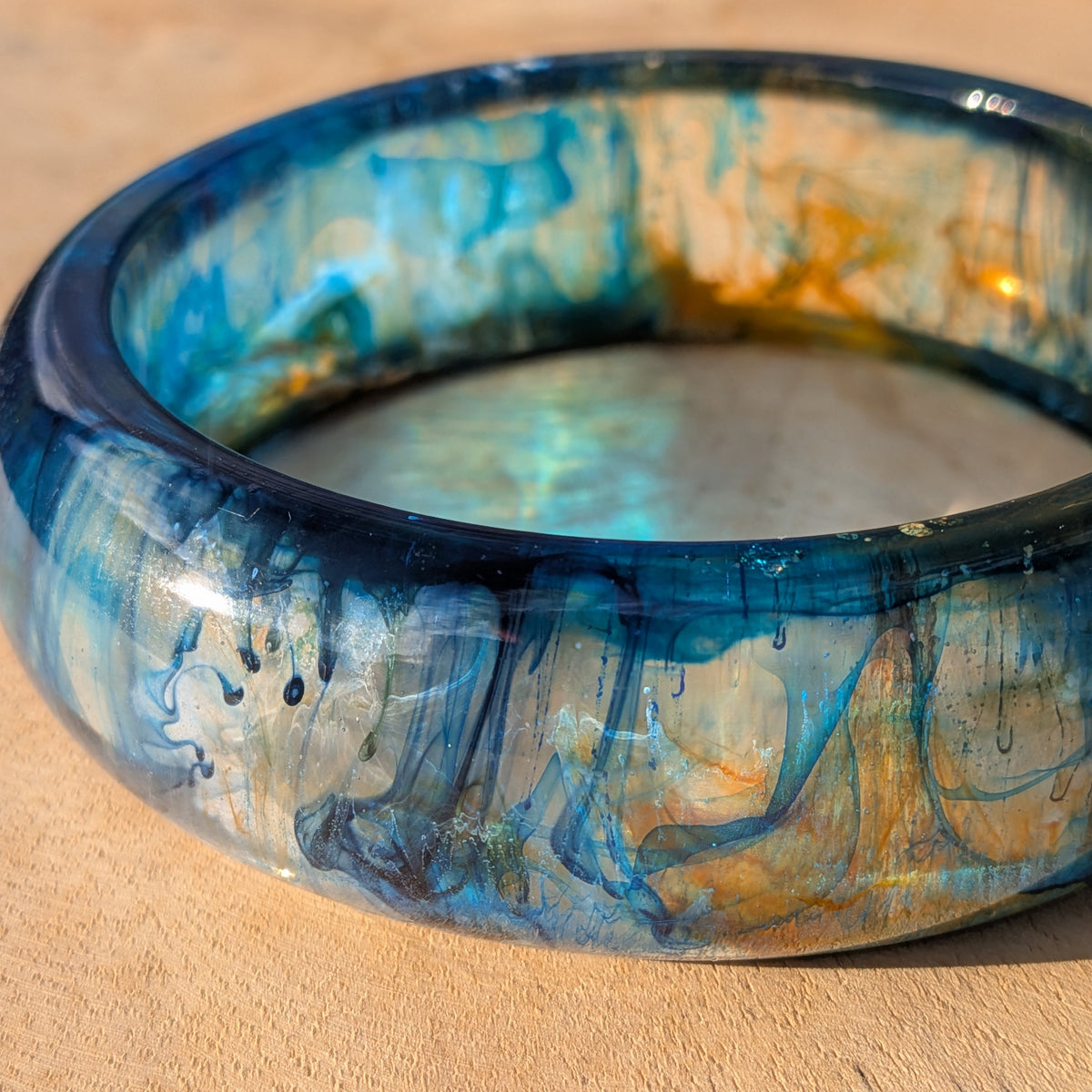 Deep blue and teal resin bangle with gold swirls lying on wooden surface – EL-Creations