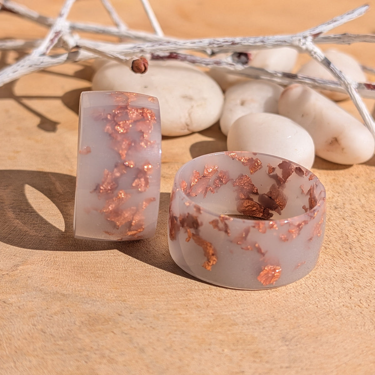 Two handmade milky lilac resin rings with copper flakes displayed together, one upright and one flat, beside white pebbles and decorative branches, EL-Creations