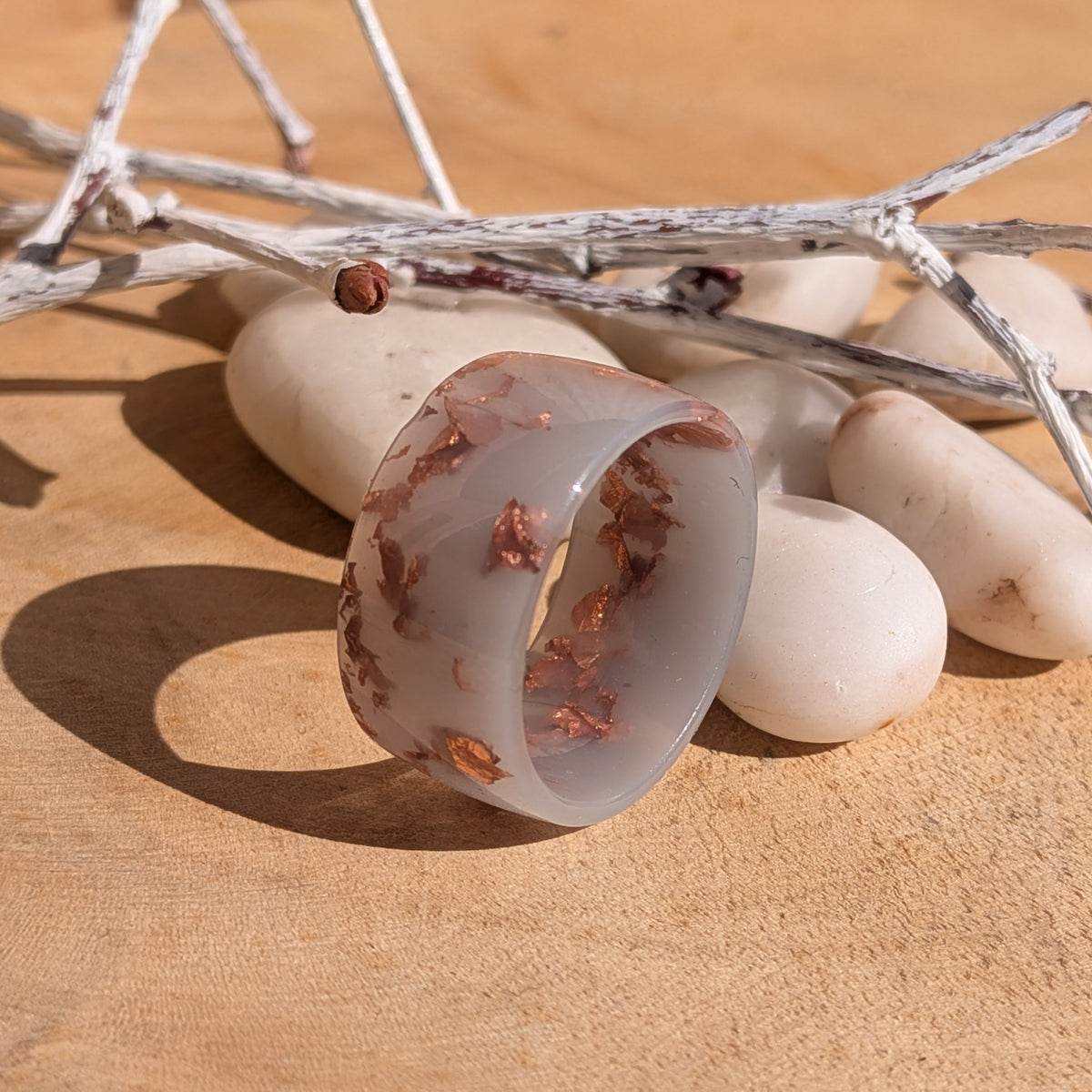 Angled view of a milky lilac resin wide-band ring with copper flakes beside white stones and silver-coloured decorative branches, EL-Creations