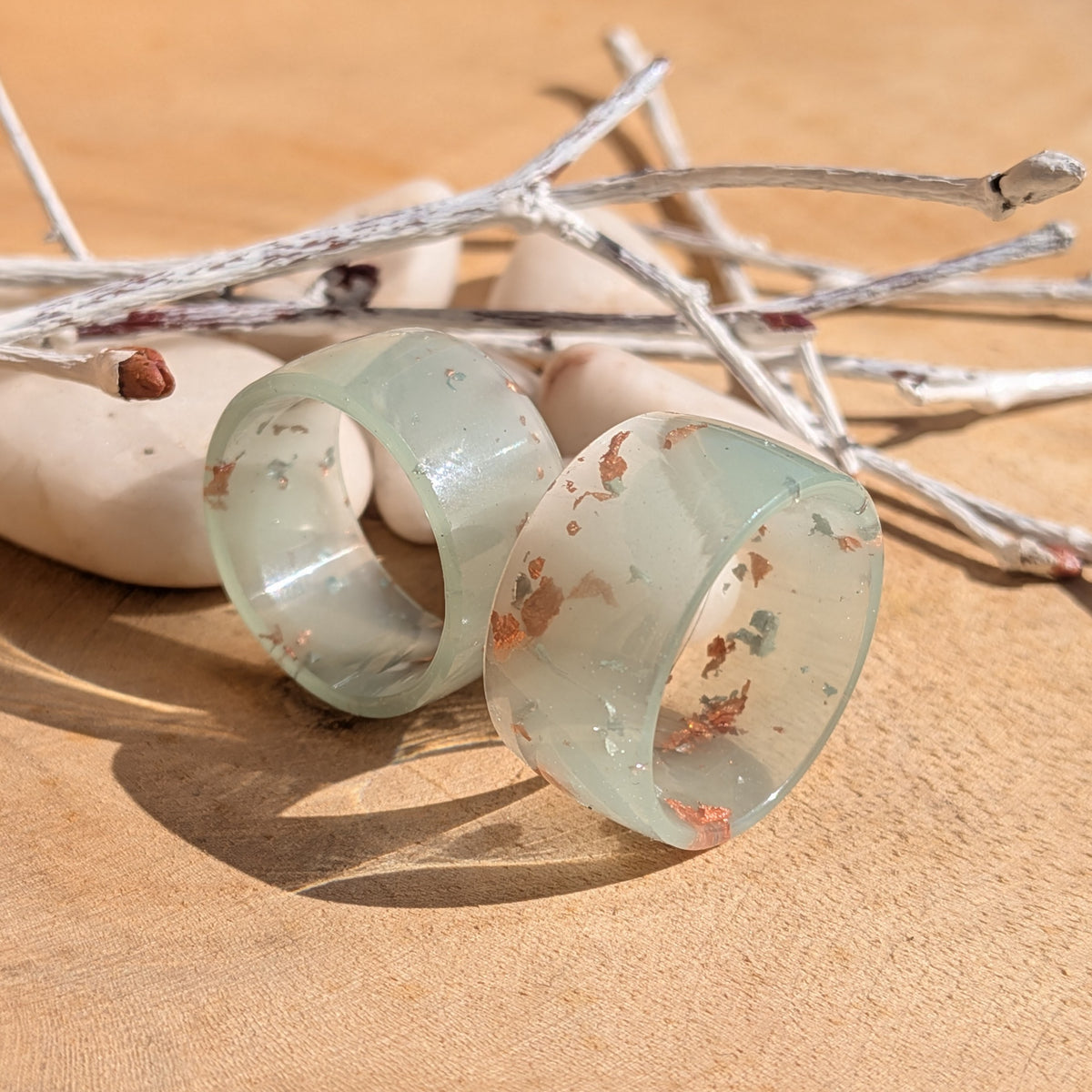 Two handmade milky blue-green resin rings with silver and copper flakes displayed beside white pebbles and decorative branches, EL-Creations