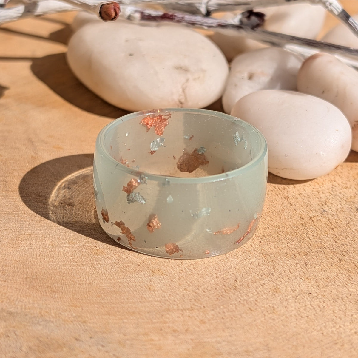 Milky blue-green resin wide-band ring with silver and copper flakes on wood in sunlight, with white pebbles behind it, EL-Creations