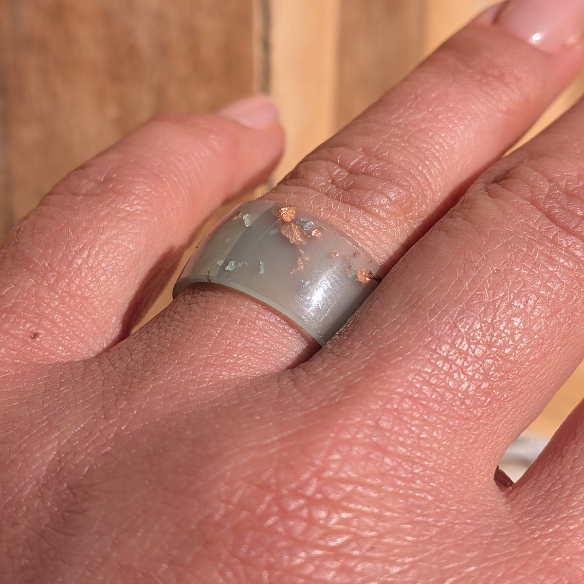 Angled close-up of a milky blue-green resin wide-band ring with silver and copper flakes worn on a finger in sunlight, EL-Creations