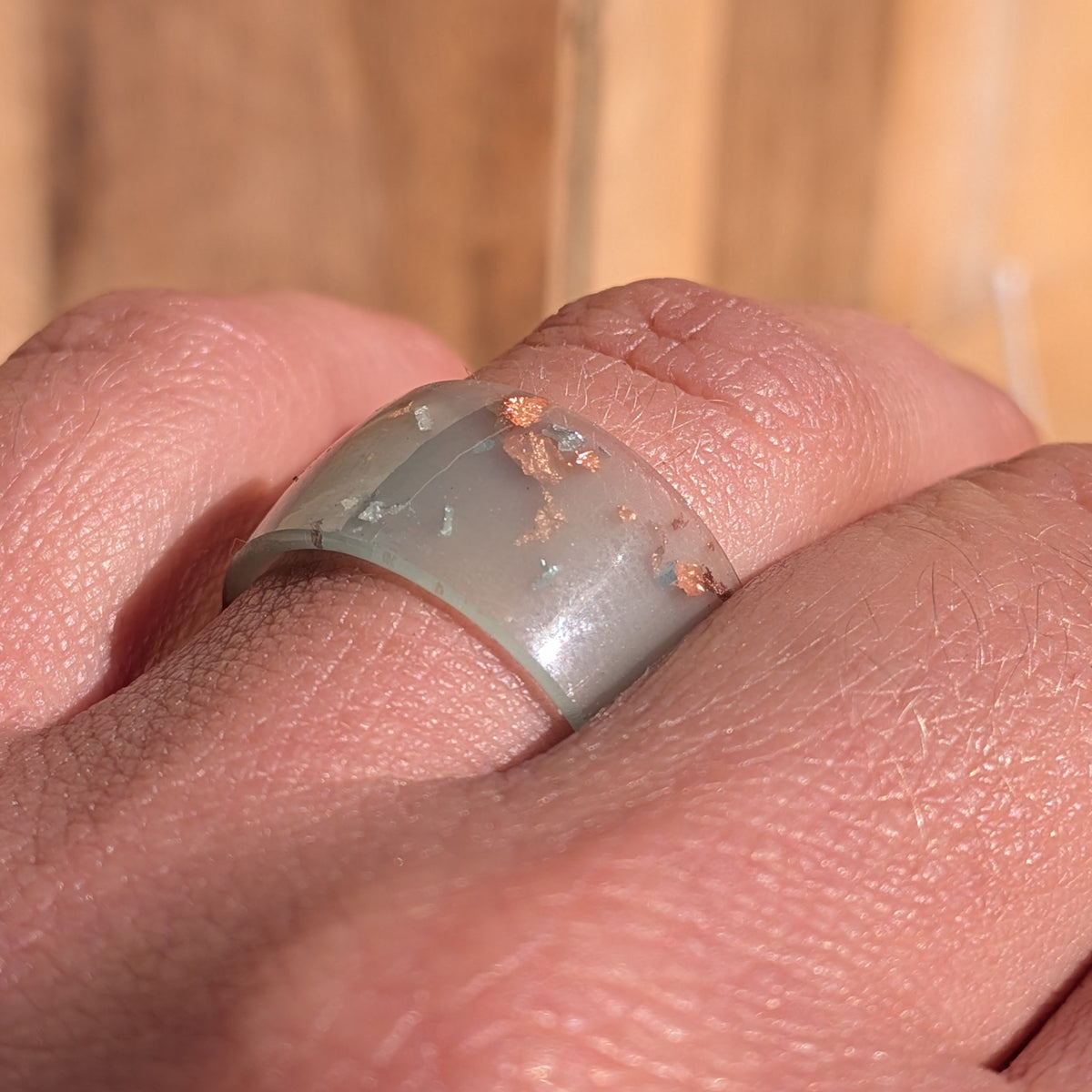 Close-up of a glossy milky blue-green resin ring worn on a finger, showing silver and copper flakes beneath the surface, EL-Creations