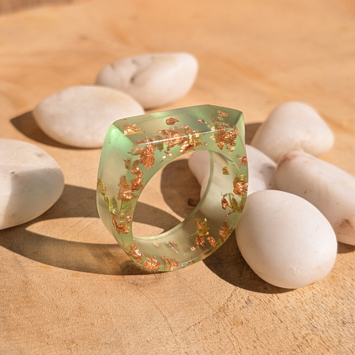 Geometric green resin ring with copper and gold flakes standing at an angle beside white pebbles on wood, EL-Creations