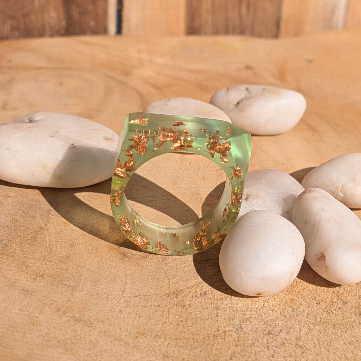 Front view of a green resin ring with copper and gold flakes standing upright among white stones in sunlight, EL-Creations