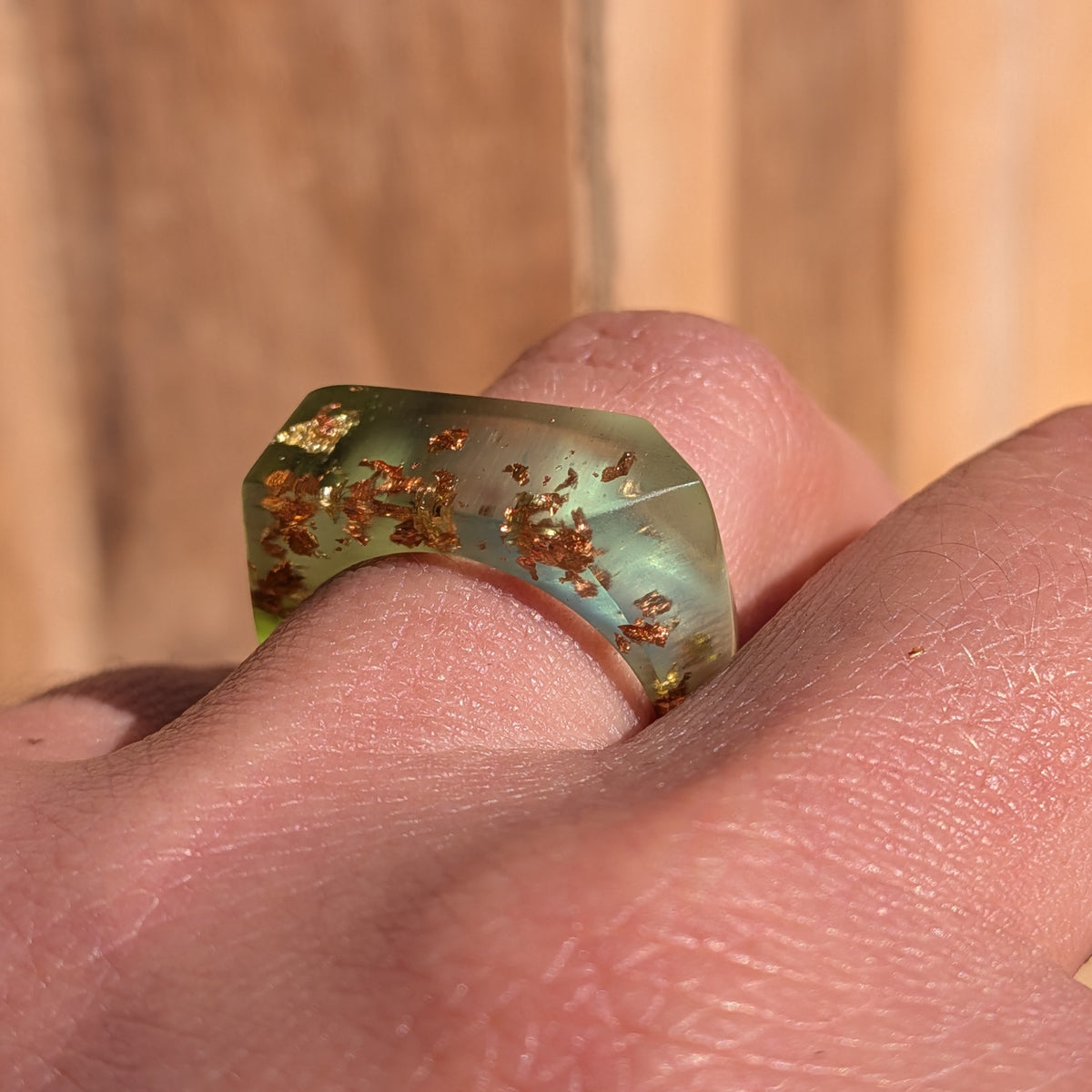 Close-up of a geometric green resin ring with copper and gold flakes worn on a finger in sunlight, EL-Creations