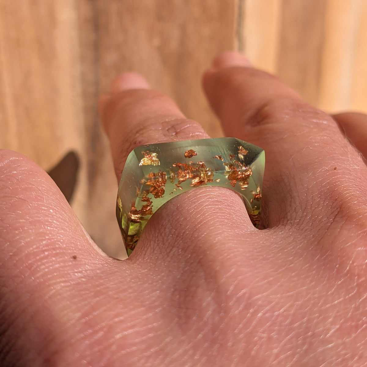 Low angled view of a green resin ring worn on a finger, showing its faceted shape and copper and gold flakes, EL-Creations