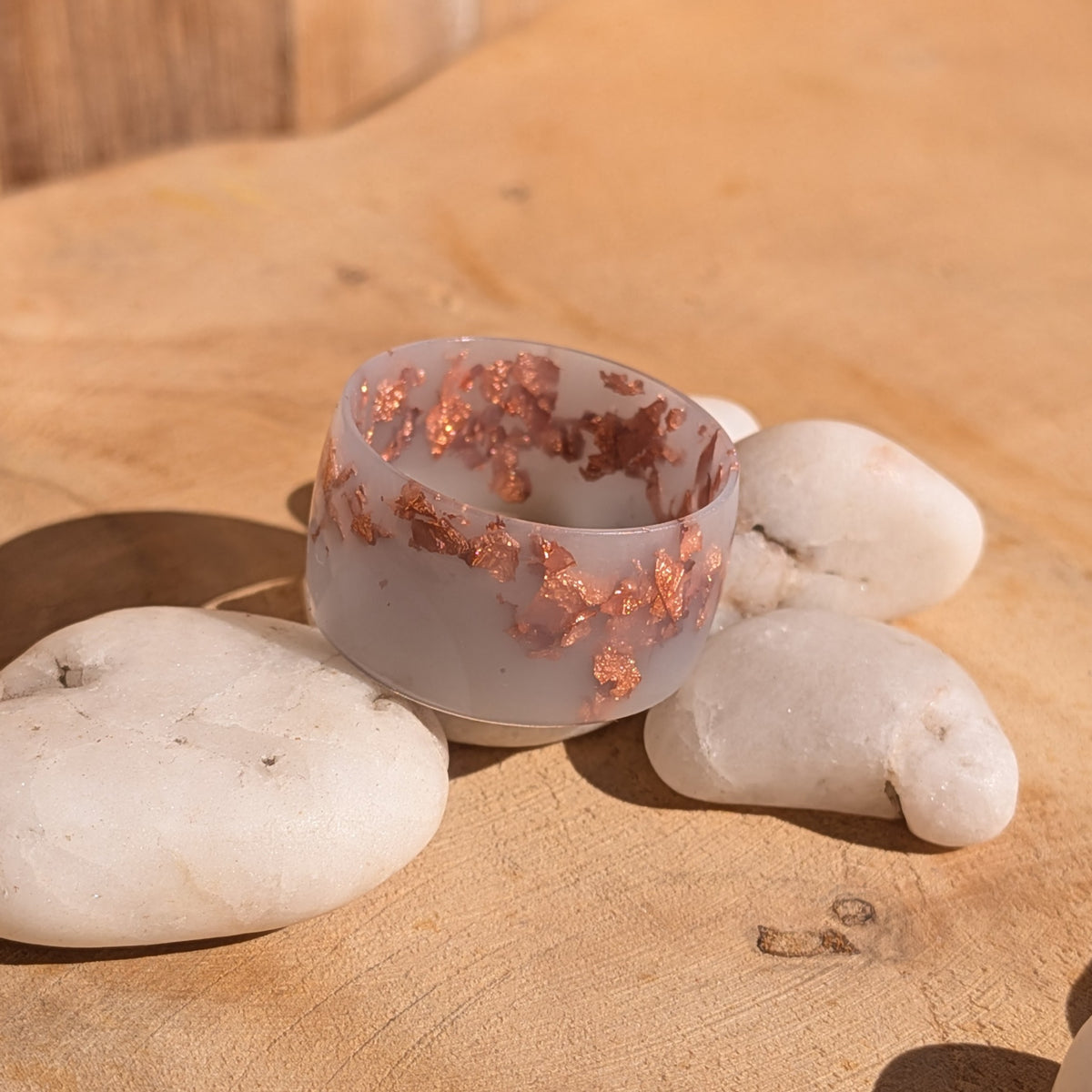 Milky lilac resin ring with copper flakes resting among white pebbles, showing its glossy wide band and inner opening, EL-Creations