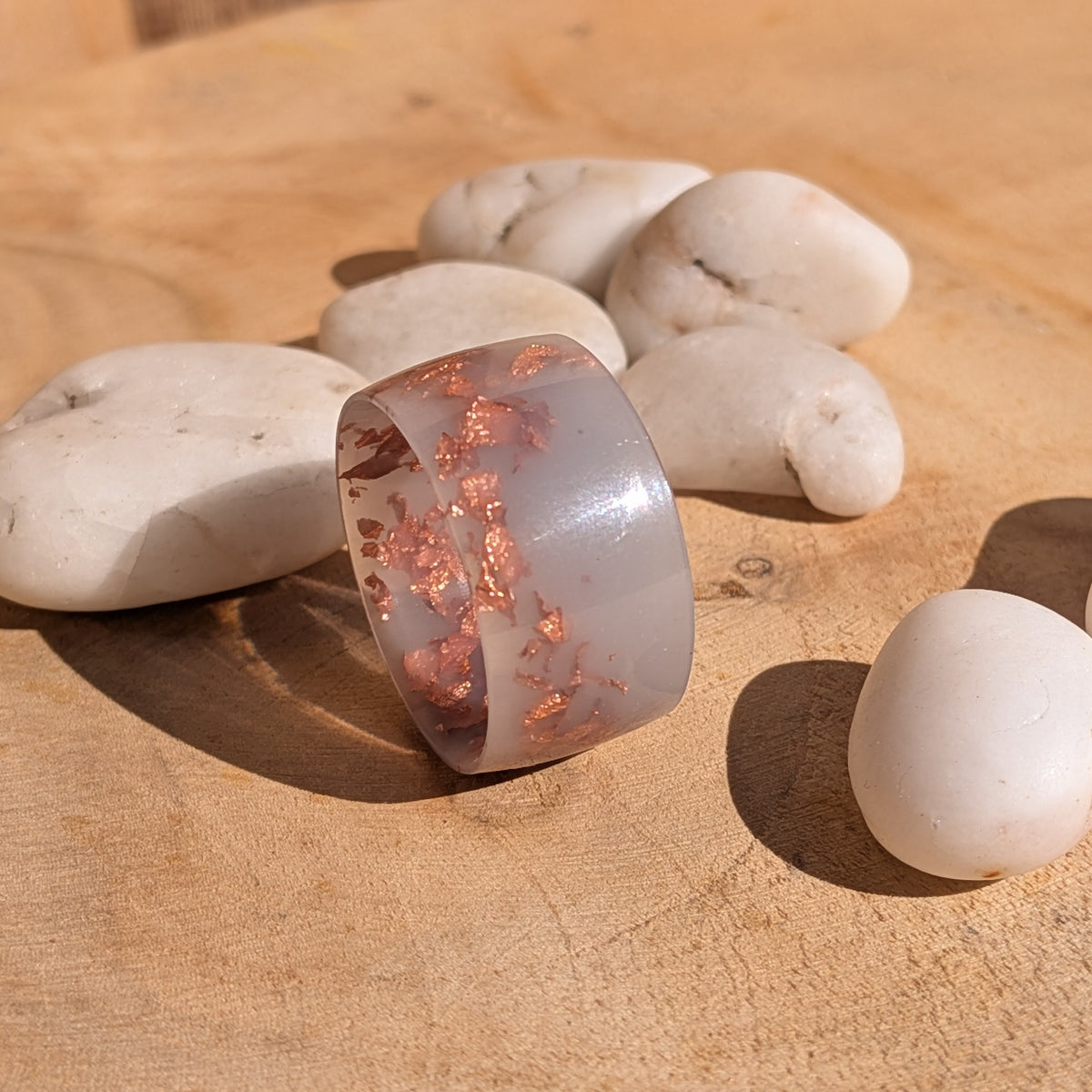 Milky lilac copper flake resin ring standing on its edge on a wooden surface beside white stones in sunlight, EL-Creations