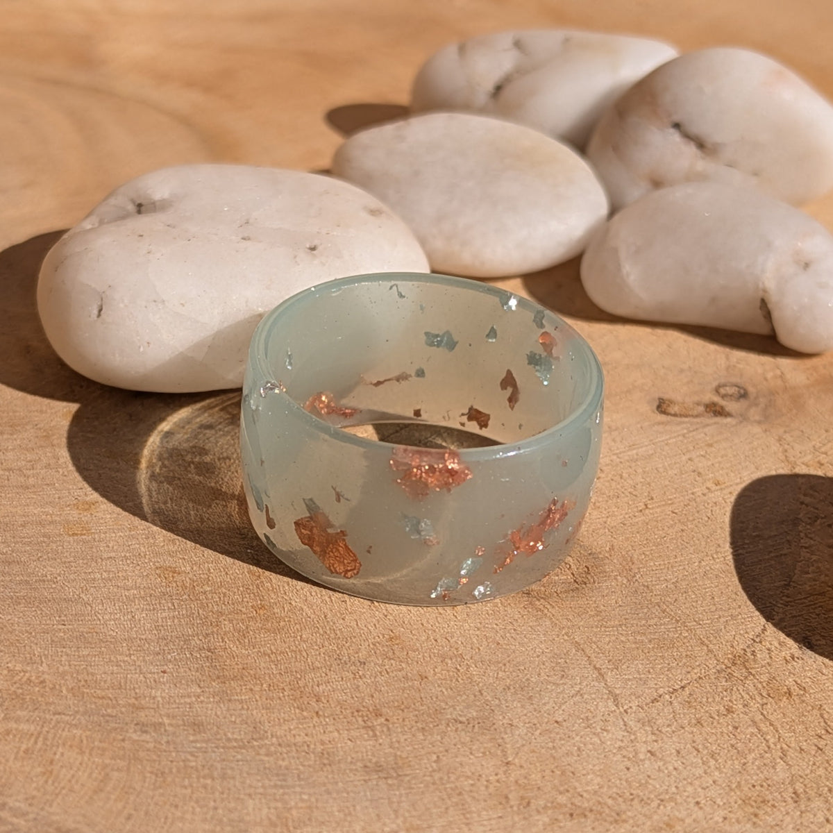 Angled view of a milky blue-green resin ring with silver and copper flakes on a wooden surface beside white stones, EL-Creations