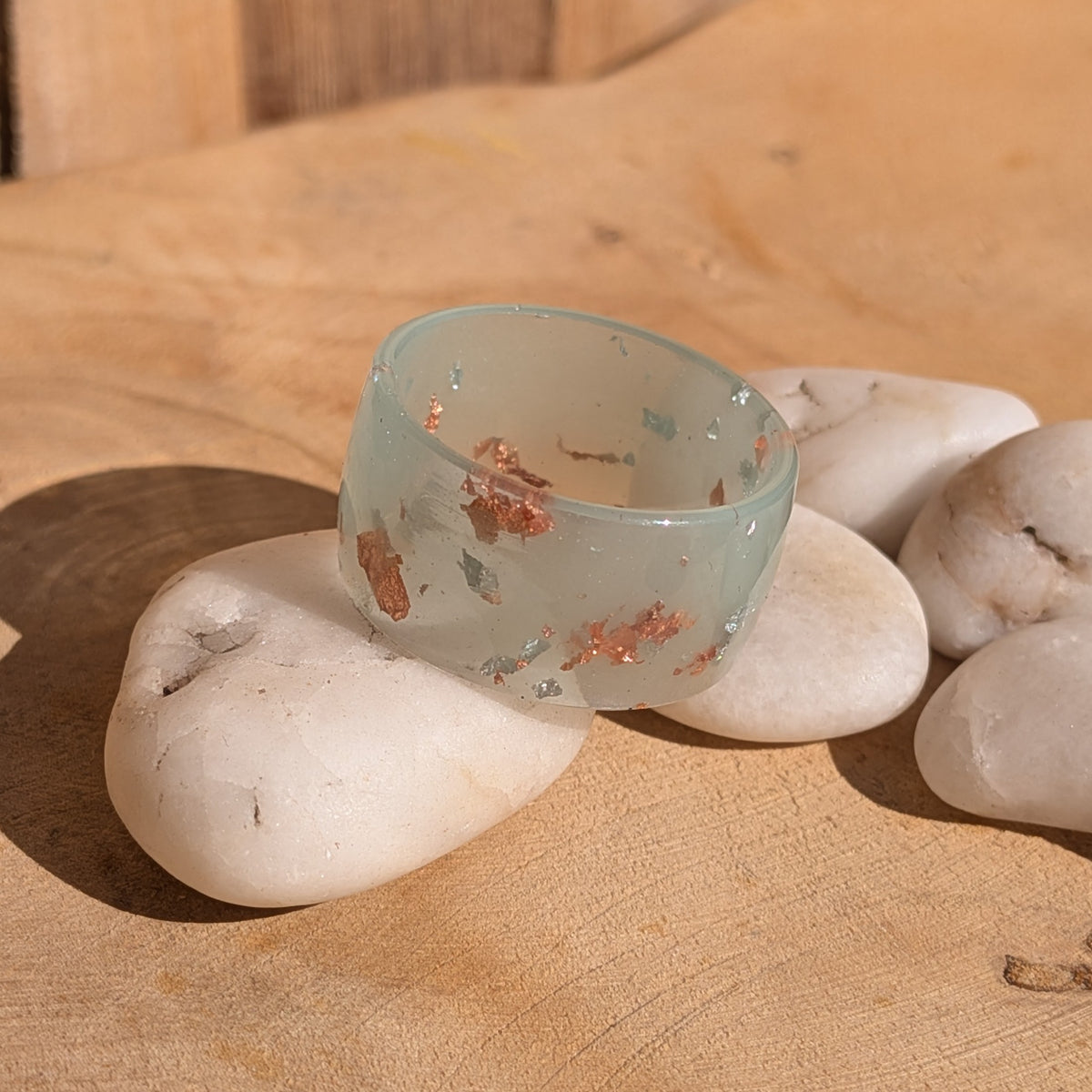 Milky blue-green resin ring with silver and copper flakes resting among white pebbles, showing its glossy band and inner opening, EL-Creations