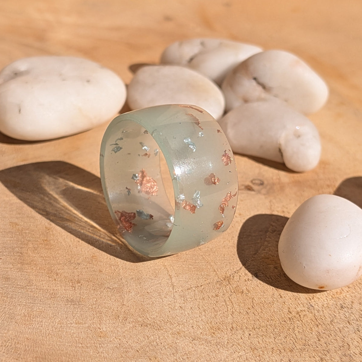 Milky blue-green resin ring with silver and copper flakes standing on its edge on a wooden surface beside white pebbles, EL-Creations