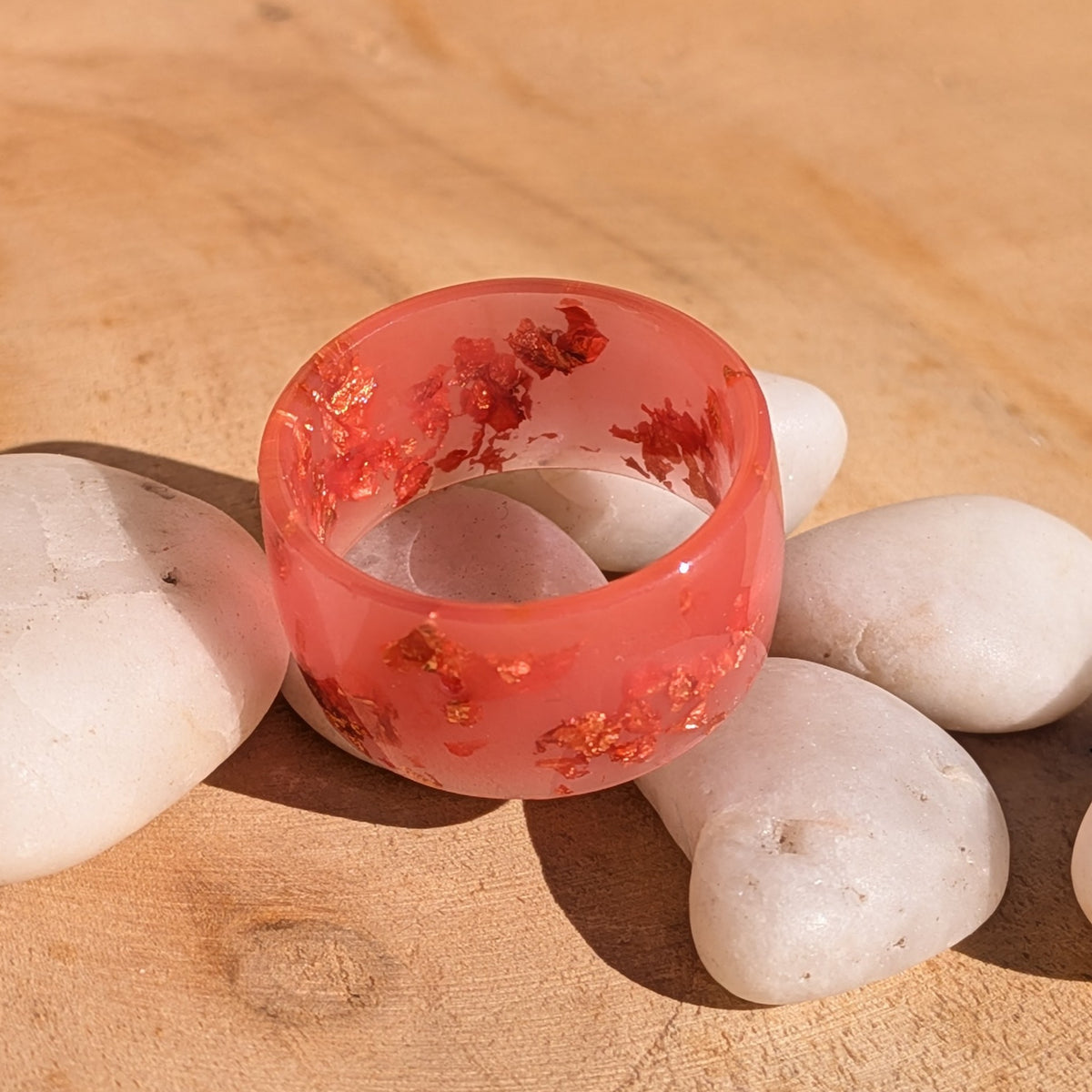 Milky pink resin ring with copper flakes nestled among white pebbles, showing its glossy wide band and inner opening, EL-Creations