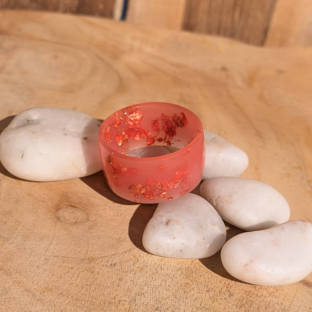 Wide milky pink resin ring with copper flakes surrounded by white pebbles on a wooden surface in sunlight, EL-Creations