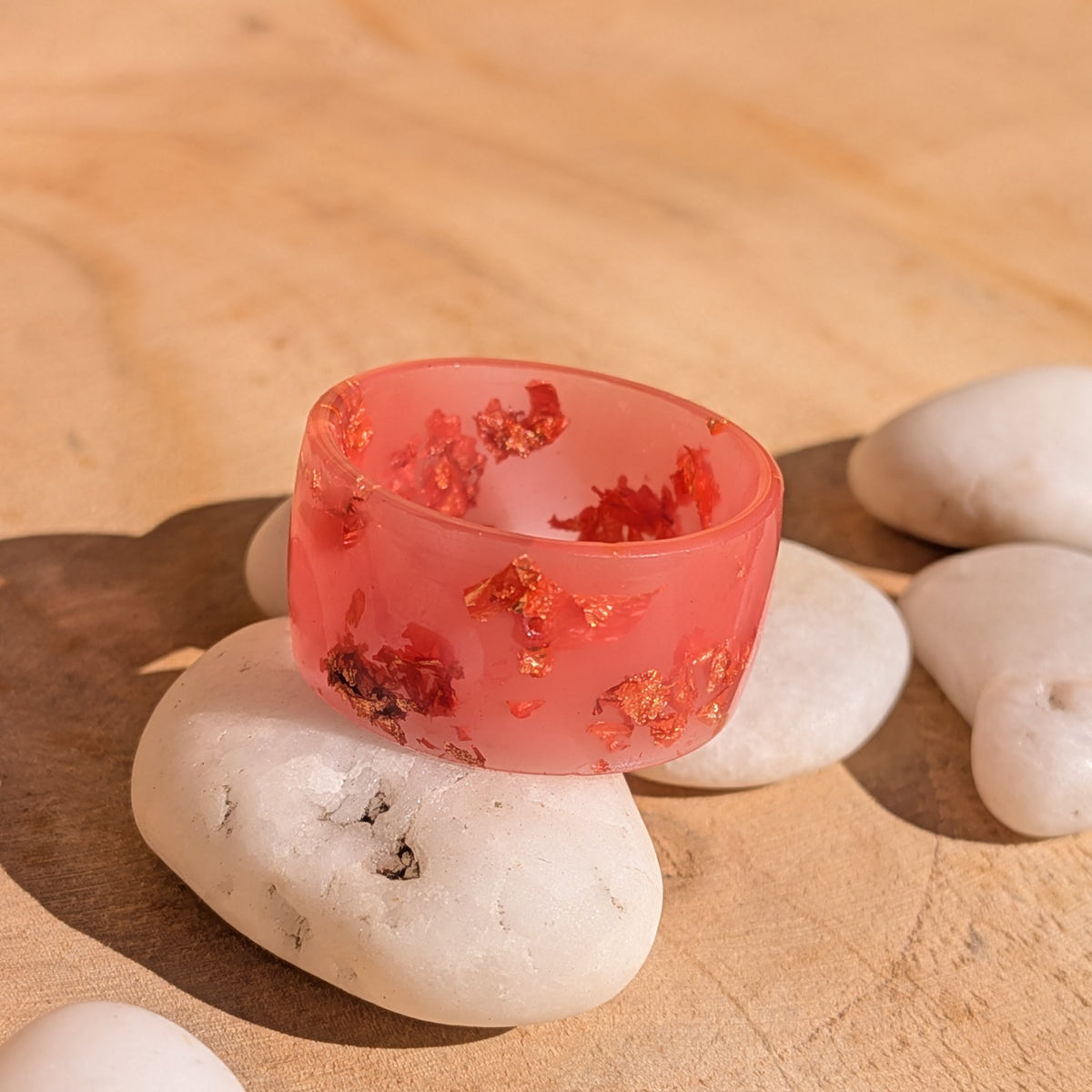 Handmade milky pink copper flake resin ring resting on a white pebble, with metallic details across the front of the band, EL-Creations