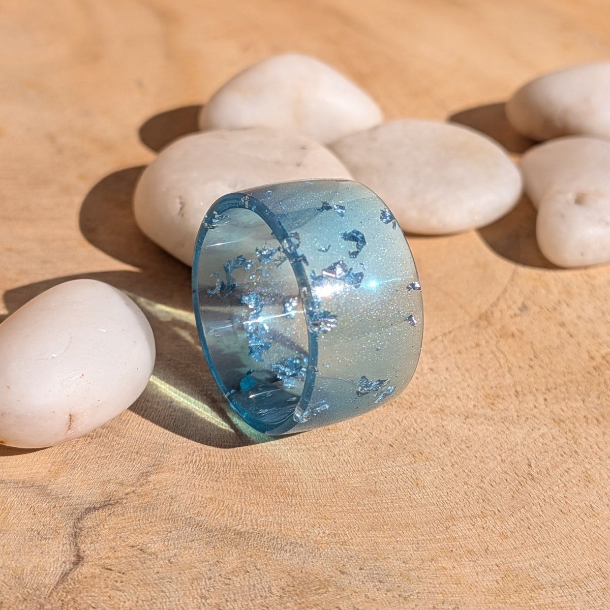 Handmade blue resin ring with silver-coloured flakes standing on a wooden surface, with sunlight reflecting across its glossy band, EL-Creations