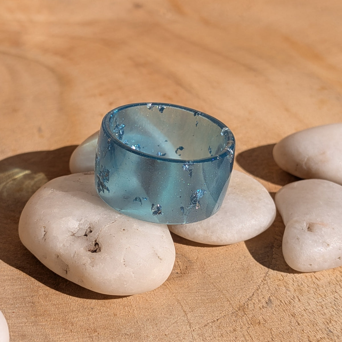 Blue resin wide-band ring resting on white pebbles, showing scattered silver-coloured flakes and its inner opening, EL-Creations