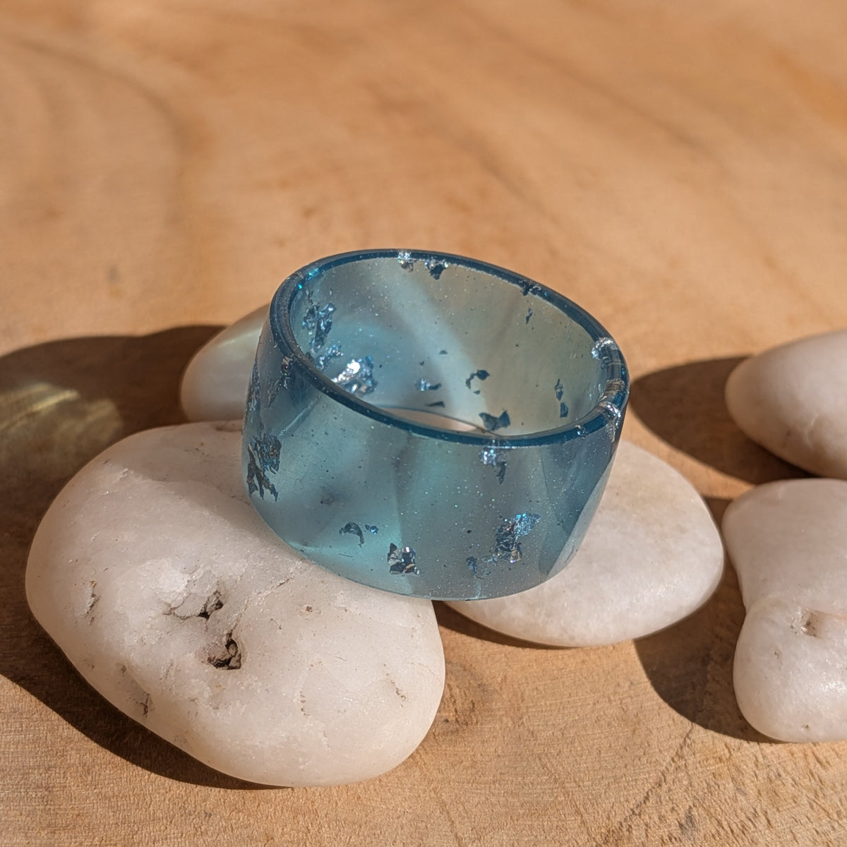 Close-up of a glossy blue resin ring with silver-coloured flakes resting among white stones in sunlight, EL-Creations