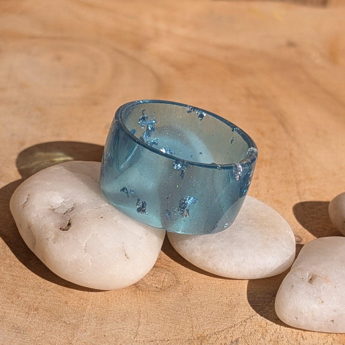 Angled view of a translucent blue resin statement ring balanced on white pebbles, with silver-coloured flakes visible inside the band, EL-Creations