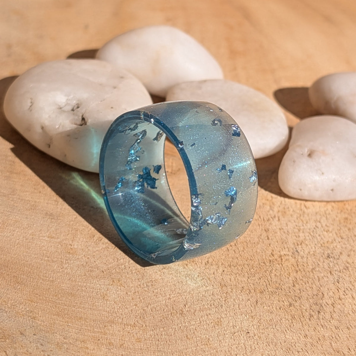 Blue resin ring with silver-coloured flakes standing upright beside white pebbles, showing its translucent wide band and circular opening, EL-Creations