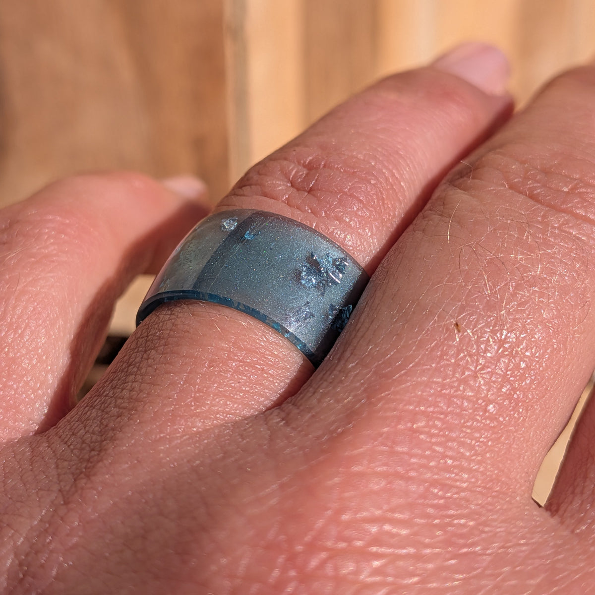 Angled close-up of a blue resin ring with silver-coloured flakes worn on a finger in sunlight, EL-Creations