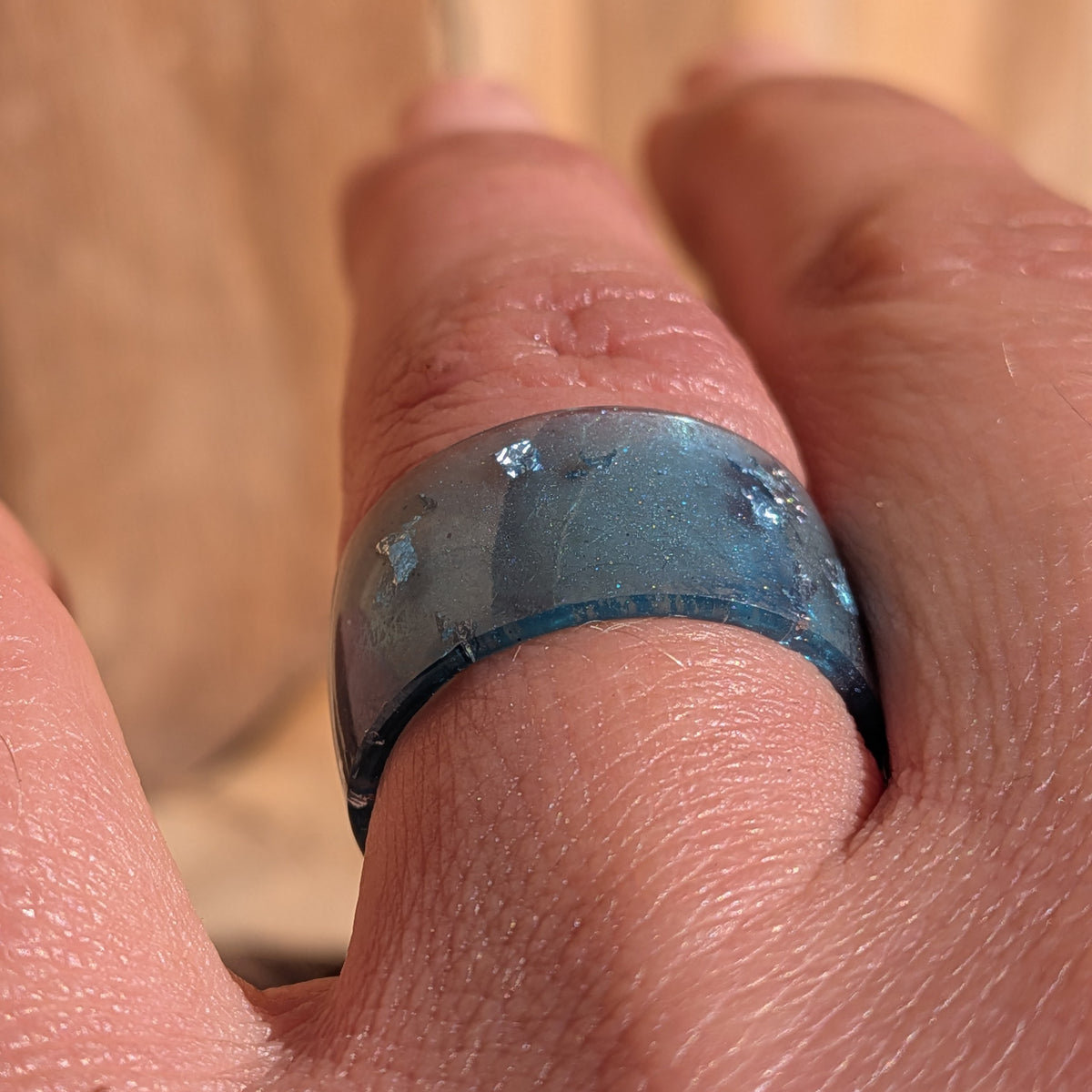Macro front view of a blue resin wide-band ring worn on a finger, showing embedded silver-coloured flakes and subtle shimmer, EL-Creations