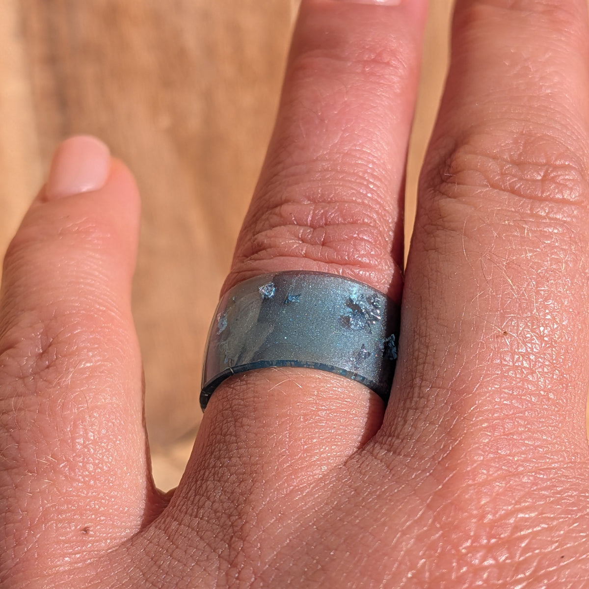 Front view of an 11 mm wide blue resin ring with silver-coloured flakes worn on a hand against a wooden background, EL-Creations