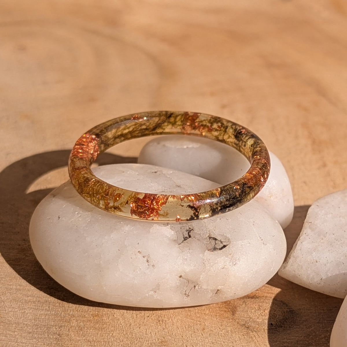 Thin resin ring with moss and copper flakes displayed on white pebbles in sunlight, EL-Creations