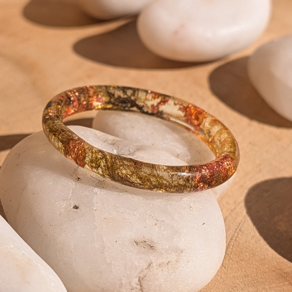 Angled close-up of a slender resin ring with moss and copper flakes on a white stone, showing its glossy rounded band, EL-Creations