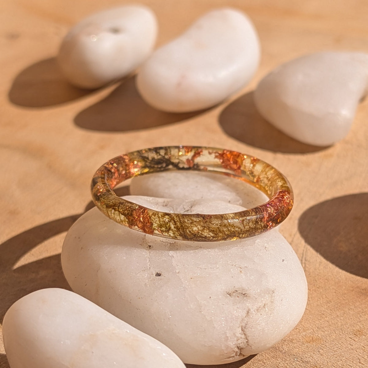 Delicate moss and copper flake resin ring resting on a white pebble with surrounding stones on a wooden surface, EL-Creations