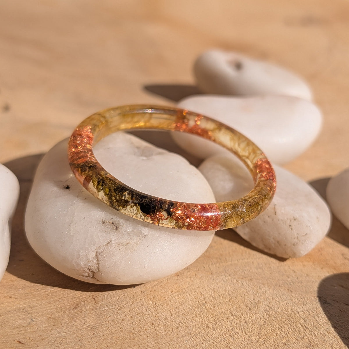 Angled view of a thin moss resin ring with copper flakes resting across white pebbles in sunlight, EL-Creations
